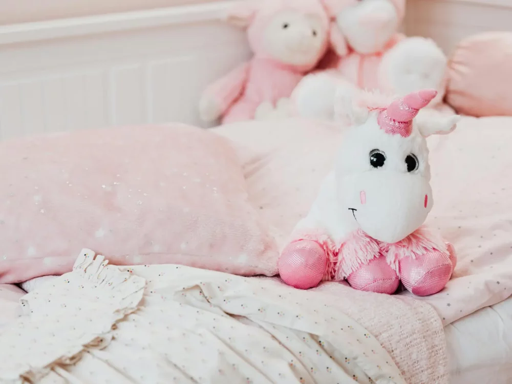 A soft unicorn plush toy sits on a pastel pink bed with pillows and blankets in a child's room.